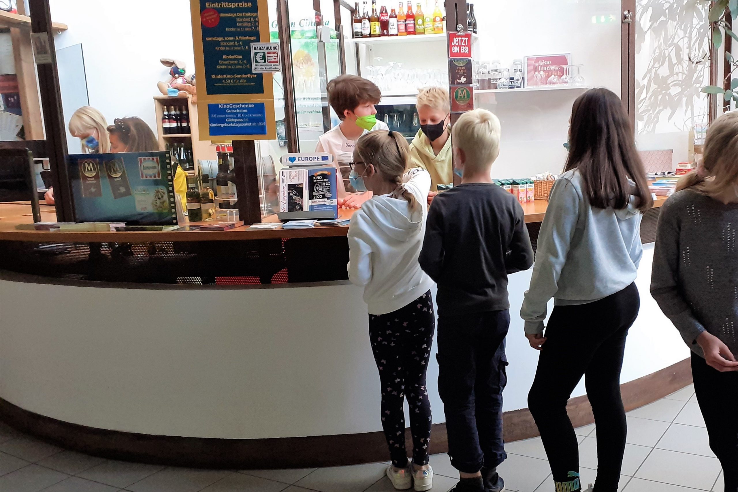 Mehrere Kinder stehen in einer Schlange vor einer Theke, an der Getränke und Snacks verkauft werden. Zwei Kinder stehen hinter der Theke.