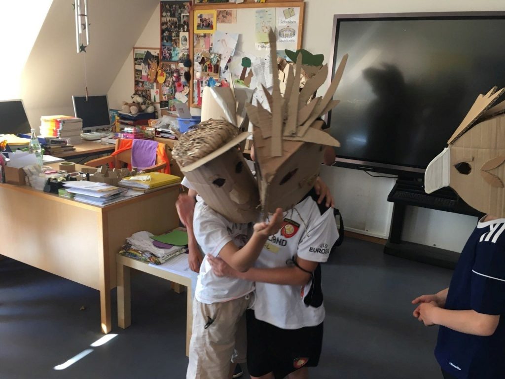 Zwei Kinder, die eine Maske aus Karton tragen stehen in einem Klassenraum und umarmen sich.