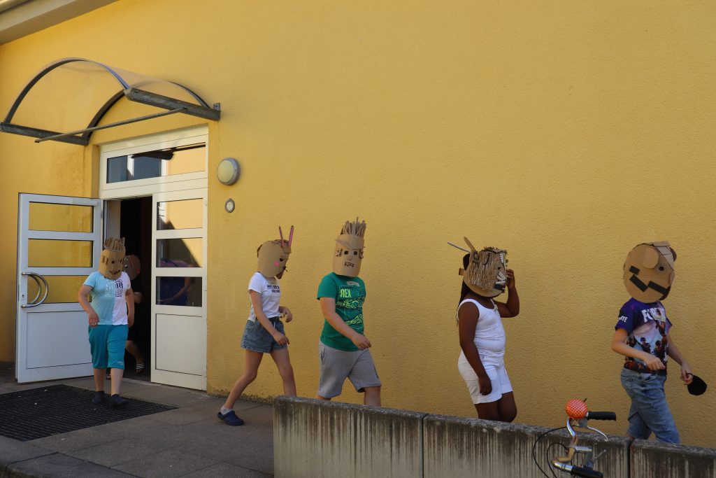 Kinder, die gebastelte Masken aus Karton tragen gehen hintereinander aus der Tür eines gelben Gebäude.