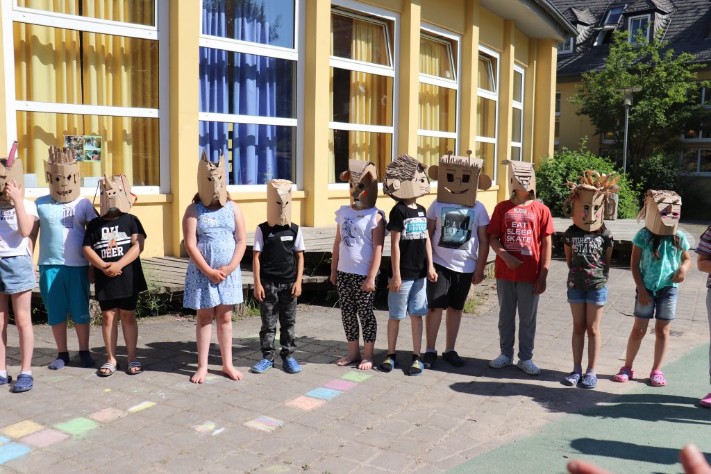 Kinder, die gebastelte Masken aus Karton tragen, stehen in einer Reihe vor einem gelben Gebäude.