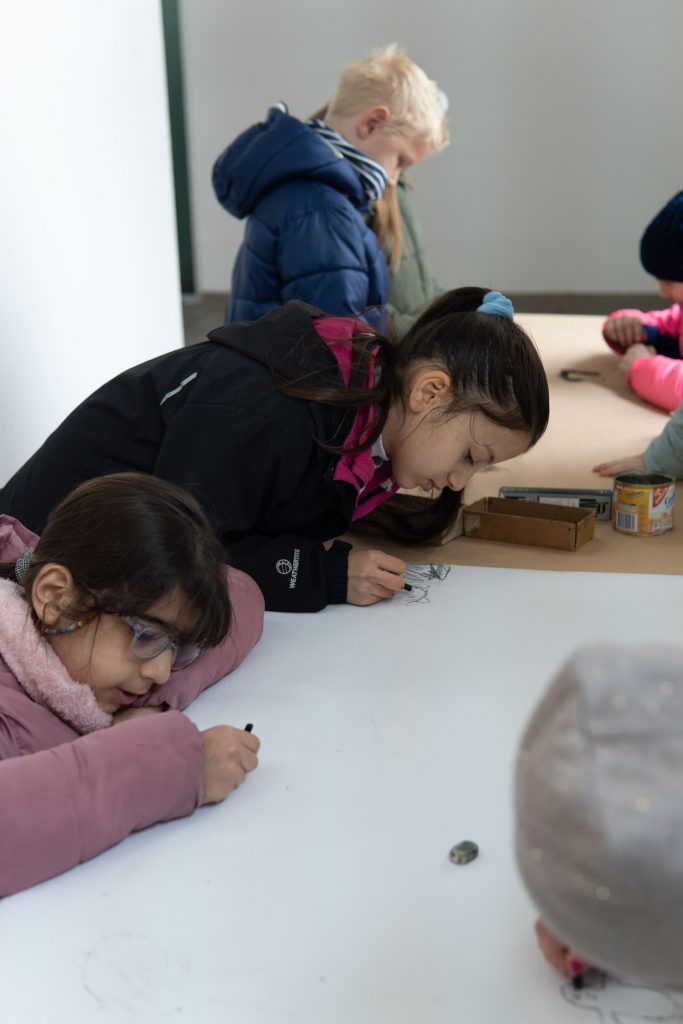 Mehrere Kinder stehen an einem Tisch und zeichnen auf einem großen weißen Blatt Papier.