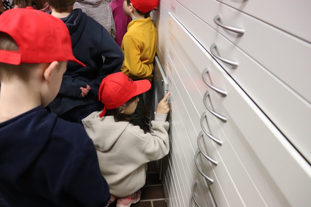 Ein Kind mit einer roten Kappe hockt vor einem Schrank mit Schubladen und zeigt auf ein Schild auf einer der Schubladen.